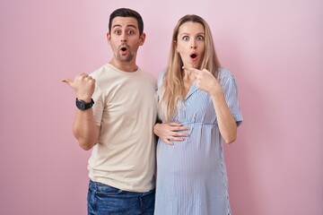 Young couple expecting a baby standing over pink background surprised pointing with finger to the...