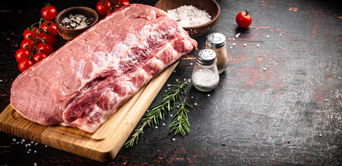 A piece of raw pork on a cutting board with tomatoes, spices and rosemary.