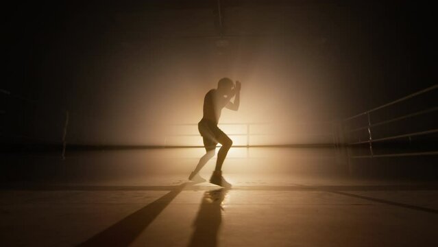 Silhouette of strong, active man exercising in dark boxing club alone. Close-up view of unrecognized, young boxer training indoors. High quality 4k footage in golden orange bronze foggy back light