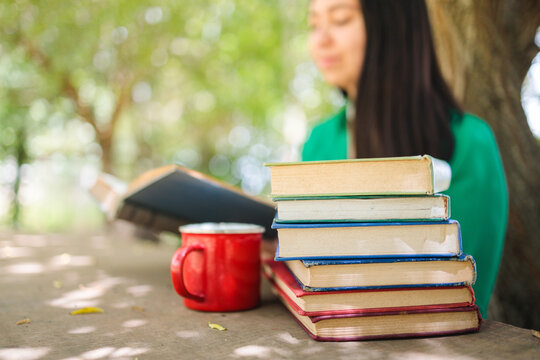 Defocused young woman reading stacked books in the field under a willow tree. Selective focus