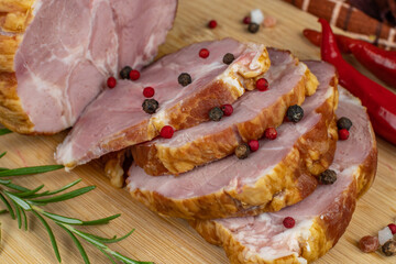 Sliced slices of juicy pork ham on a wooden cutting board, next to it are a carving knife, multi-colored allspice peas, red hot peppers and basil. Close-up. The concept of delicious meat products