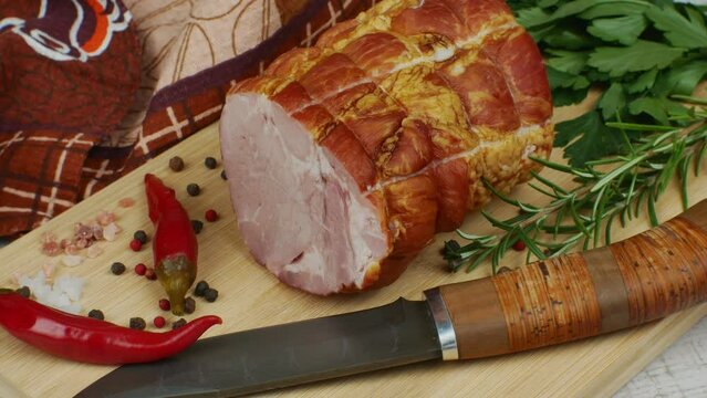 On a wooden cutting board, a juicy piece of pork ham, a carving knife, red hot peppers, multi-colored allspice, parsley, dill and basil. The concept of delicious pork products