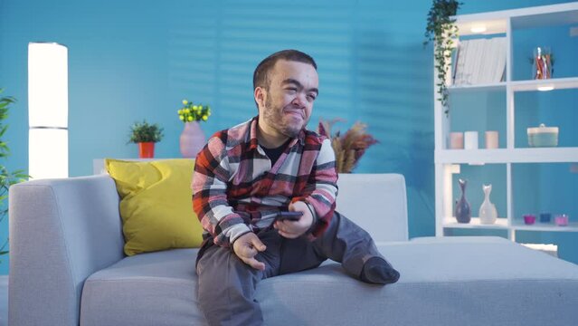 Young dwarf man guy holding smartphone device sending sms message at home, playing mobile game, disabled man using apps chatting on social media.