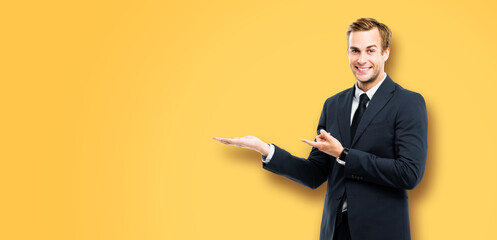 Businessman in black suit showing giving advertising some product, on vivid yellow background. Copy...