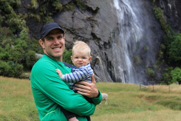 father holding baby in front of a waterfall in new zealand