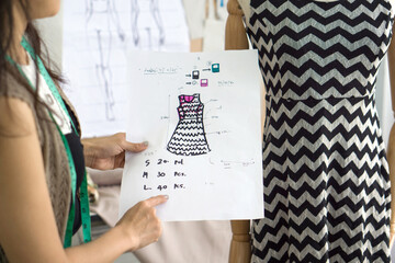 Young fashion design business owner checking orders from note on pattern for cloth, comparing with the dress on the mannequin.