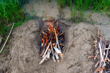 A bonfire made of birch branches taken from above in the sand next to grass and firewood.