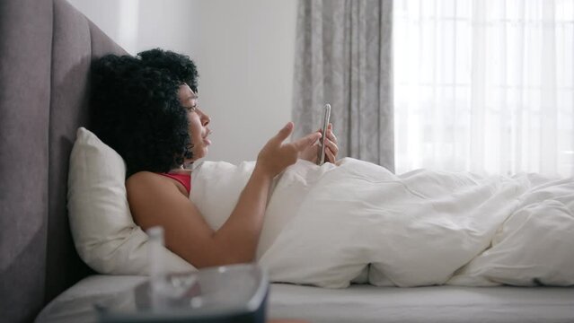 Smiling girl on video call with friends or family. Slow motion wireless connection and communication. Side view bedroom apartment, beautiful African American woman using smartphone in bed at morning