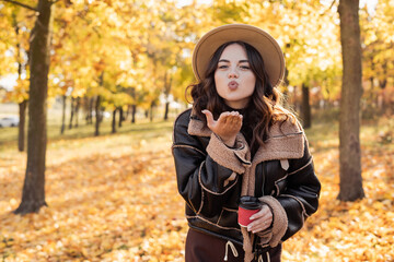 Autumn portrait stylish woman blowing lips sending air kiss in the city park on yellow leaves background.