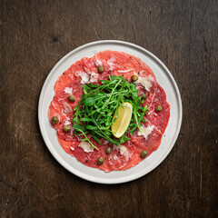 Beef carpaccio with capers, arugula and parmesan on a white plate on a dark wooden background