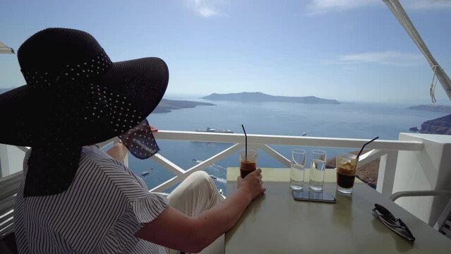 Happy woman drinking frappe coffee enjoying summer vacation at luxury hotel cafe with amazing landscape view of Fira, Santorini. Europe travel destination. Tourist lifestyle.