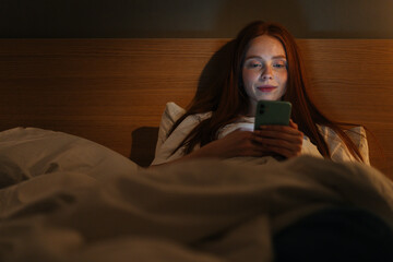 Portrait of happy smiling young woman using smartphone, looking on screen, typing online message on social media, lying on bed late at night, bedside lamp lighting with warm yellow light.
