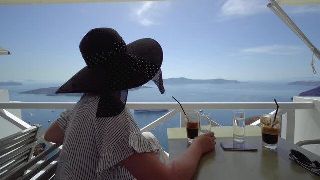 Breakfast woman sitting at luxury travel restaurant terrace drinking frappe coffee cup over Mediterranean view from outside balcony. Greek healthy food Santonini vacation.