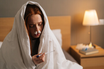 Close-up face of sick tired redhead woman suffering from runny nose disease with paper tissues into nose, measuring temperature with thermometer, sitting in bed covered with warm blanket at home.