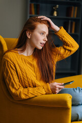 Vertical portrait of attractive redhead woman using mobile phone sitting in yellow armchair,...