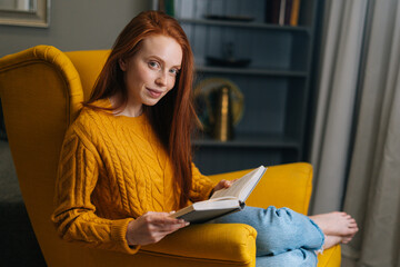 Portrait of elegance redhead woman reading exciting story having relaxing weekend sitting in yellow armchair, smiling looking at camera. Beautiful female relaxing enjoying lazy morning at home.