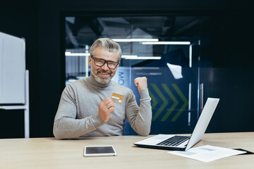 Successful gray haired businessman rejoices and celebrates completion of money transfer transaction, portrait of boss inside office with bank credit card and laptop, mature man looking at camera.