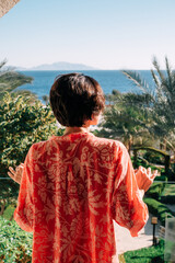 Shot of  woman with short hair in red tunic standing on the balcony and looking at the sea view. Vacation summer travel