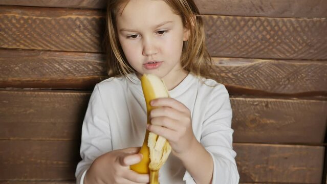 Cute cute teen girl peels a big ripe yellow banana and eats it. Portrait of a girl biting off a banana on the background of a brown wall of boards. healthy eating fruits