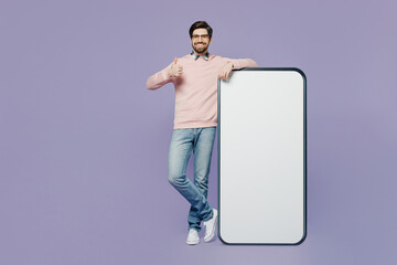 Full body happy young IT man he wears casual clothes pink sweater glasses big huge blank screen mobile cell phone smartphone with area show thumb up isolated on plain pastel light purple background.