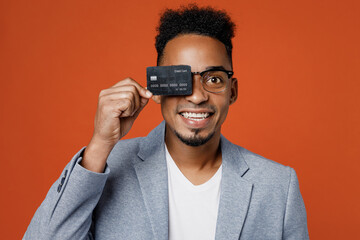 Young rich happy employee business man corporate lawyer wear classic formal grey suit shirt glasses work in office hold cover eye with credit bank card isolated on plain red orange background studio.