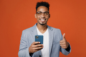 Young happy employee business man corporate lawyer wear classic formal grey suit shirt glasses work in office hold use mobile cell phone show thumb up isolated on plain red orange background studio.
