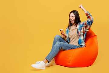 Full body young woman wear blue shirt beige t-shirt sit in bag chair hold in hand use mobile cell phone do winner gesture isolated on plain yellow background studio portrait. People lifestyle concept.