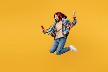 Full body side view young excited fun cheerful caucasian woman in blue shirt beige t-shirt headphones listen music use mobile cell phone jump high isolated on plain yellow background studio portrait.