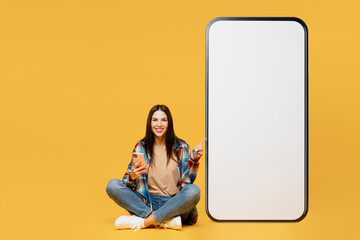 Full body fun young woman wear blue shirt beige t-shirt sit point on big huge blank screen mobile cell phone with workspace copy space mockup area use smartphone isolated on plain yellow background.