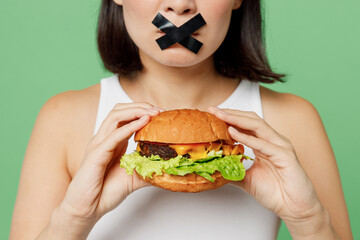 Cropped young wistful woman with mouth sealed with tape wear white clothes hold look at burger isolated on plain pastel green background. Proper nutrition healthy fast food unhealthy choice concept.