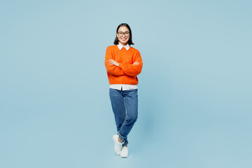 Full body young confident woman of Asian ethnicity in orange sweater glasses look camera hold hands crossed folded isolated on plain pastel light blue cyan background studio People lifestyle concept