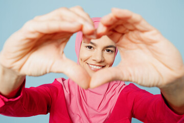 Fototapeta na wymiar Close up young arabian muslim woman wear pink abaya hijab showing shape heart look through hand heart-shape sign isolated on plain pastel light blue cyan background People uae islam religious concept