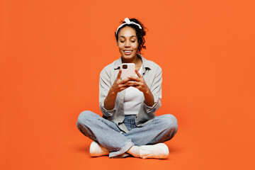 Full body fun young woman of African American ethnicity she wears grey shirt headband hold in hand use mobile cell phone isolated on plain orange background studio portrait. People lifestyle concept.