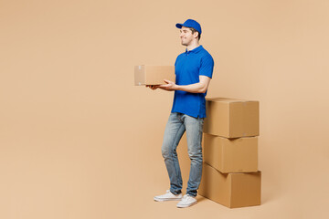 Full body sideways delivery guy employee man wear blue cap t-shirt uniform workwear work as dealer courier hold cardboard box stand near stack isolated on plain light beige background Service concept - obrazy, fototapety, plakaty