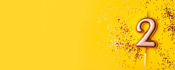 Gold candle in the form of number two on yellow background with confetti.