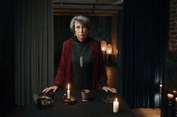 Portrait of old mature gypsy witch standing at table with psychic tools
