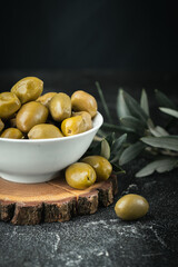 Close up shot of green olives in a white bowl on the wooden stand with olive leaves on a black background. Traditional Greek and Italian food.