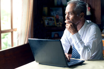 Thoughtful senior asian businessman holding hands on chin looking through window with serious doubt thinking challenging business problem sitting at desk with laptop