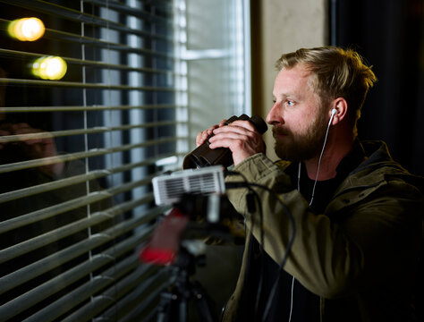 Man standing by window observing neighborhood with binoculars
