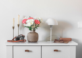 Living room interior - candles, fresh flowers in a ceramic vase, a table lamp, a notebook and a camera on a white chest of drawers in a bright Scandinavian-style room