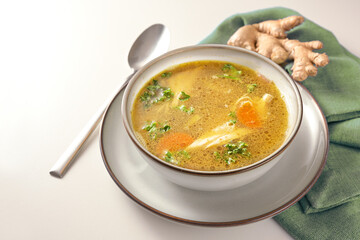 Homemade chicken soup with vegetables, ginger and parsley in a bowl, green napkin and spoon on a gay white table, copy space, selected focus