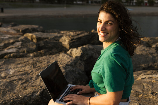 Happy woman with laptop sitting on rock at beach
