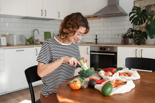 Smiling young woman cutting broccoli sitting at table in kitchen