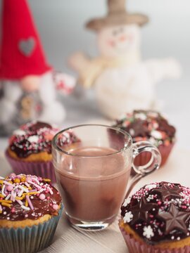 Cupcake with chocolate and colorful sugar decoration and hot chocolate in transparent cup on wooden white painted table in front of snowman