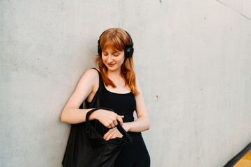 Fototapeta premium Ginger woman with headphones standing by concrete wall outdoors