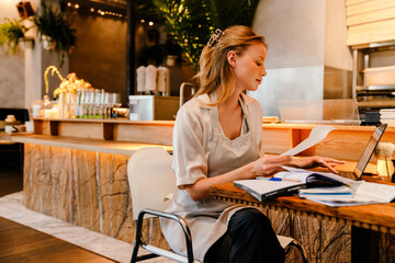 Young woman doing paperwork and using laptop while working in cafe