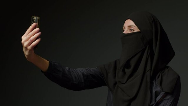 Muslim Woman Makes A Selfie Holding Mobile, Phone Point Of View, Arab Girl Wearing Hijab Smiling And Looking At The Camera On Black Background