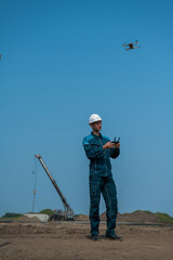 A man in a helmet and overalls controls a drone at a construction site. The builder carries out technical oversight.