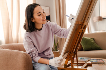 Cheerful asian woman artist drawing on canvas while sitting on armchair at home