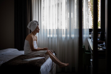 Sad young woman wrapped in towel after shower sitting on edge of bed in hotel room. Depression and...